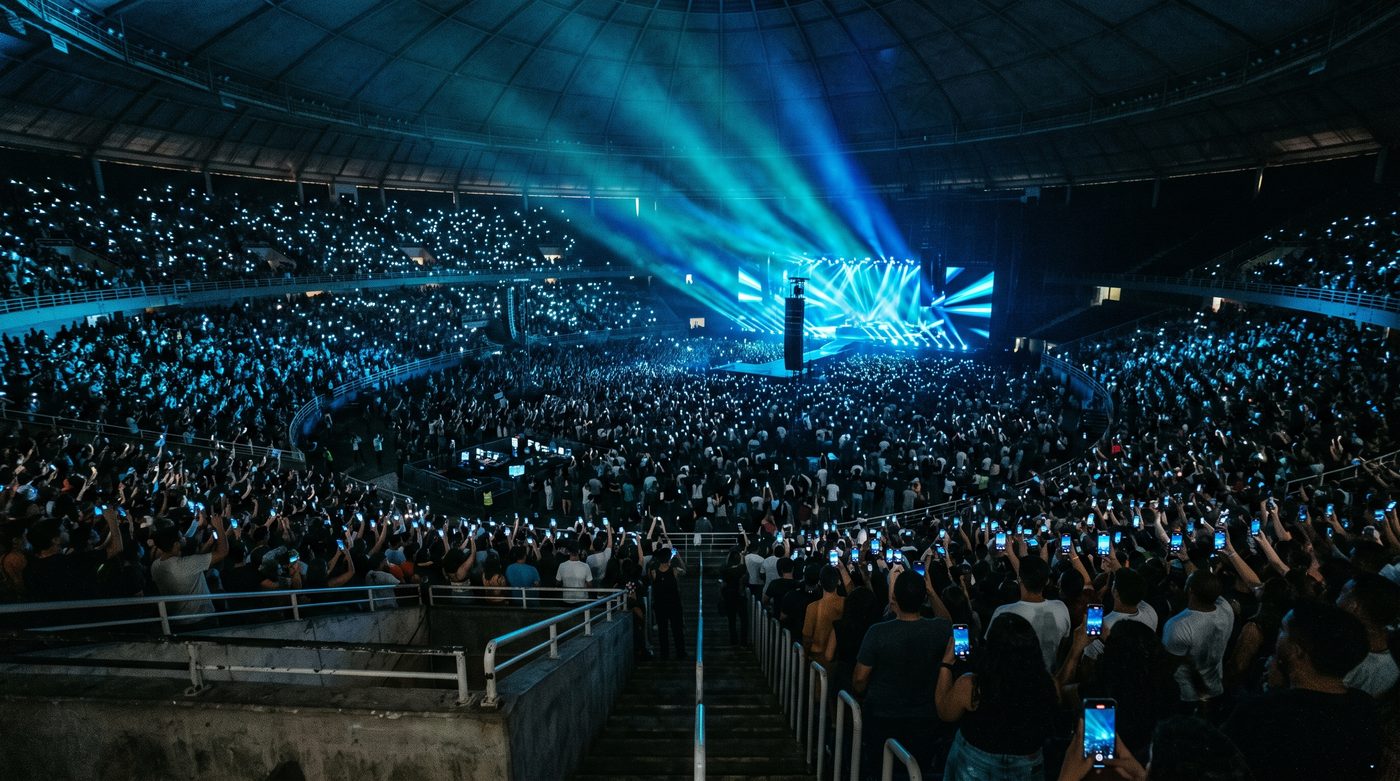 Arena lotada durante um evento noturno, com milhares de pessoas erguendo celulares iluminados diante do palco